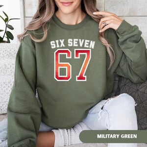 May include: An olive green sweatshirt with the text "SIX SEVEN" in white and the number "67" in red and orange. The sweatshirt is a solid color and has a crew neck. The words "MILITARY GREEN" are at the bottom of the image.