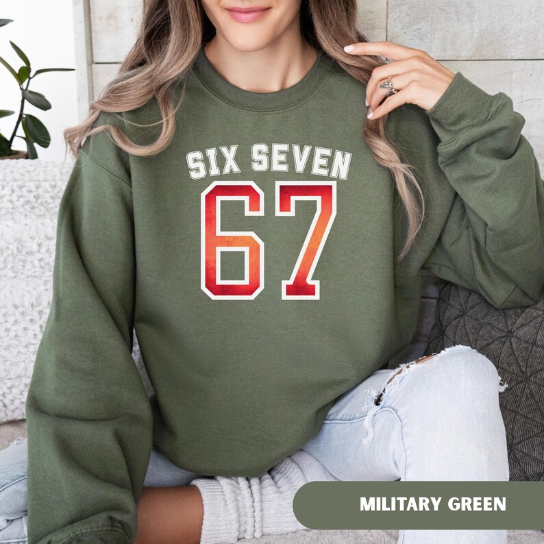 May include: An olive green sweatshirt with the text "SIX SEVEN" in white and the number "67" in red and orange. The sweatshirt is a solid color and has a crew neck. The words "MILITARY GREEN" are at the bottom of the image.