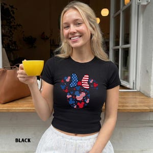 May include: Black, short-sleeved t-shirt with a patriotic Minnie Mouse design. The design uses the colours and patterns of the American flag, including stars and stripes, arranged in a bow and Mickey Mouse silhouettes. The word "BLACK" is printed at the bottom.