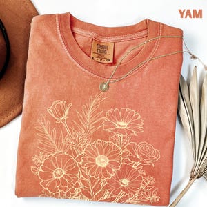 K&ouml;nnte beinhalten: Ein korallenfarbenes Comfort Colors T-Shirt mit einem cremefarbenen Blumendesign. Das T-Shirt mit Rundhalsausschnitt hat kurze &Auml;rmel. Eine goldene Halskette mit Anh&auml;nger ist &uuml;ber dem Shirt drapiert. Das Wort "YAM" befindet sich in der oberen rechten Ecke.