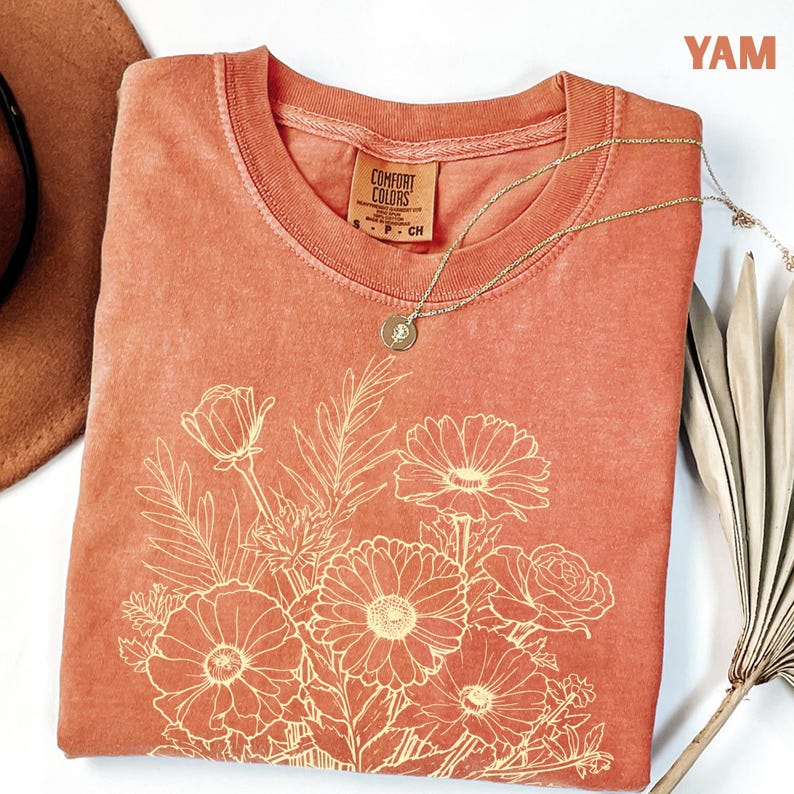 K&ouml;nnte beinhalten: Ein korallenfarbenes Comfort Colors T-Shirt mit einem cremefarbenen Blumendesign. Das T-Shirt mit Rundhalsausschnitt hat kurze &Auml;rmel. Eine goldene Halskette mit Anh&auml;nger ist &uuml;ber dem Shirt drapiert. Das Wort "YAM" befindet sich in der oberen rechten Ecke.