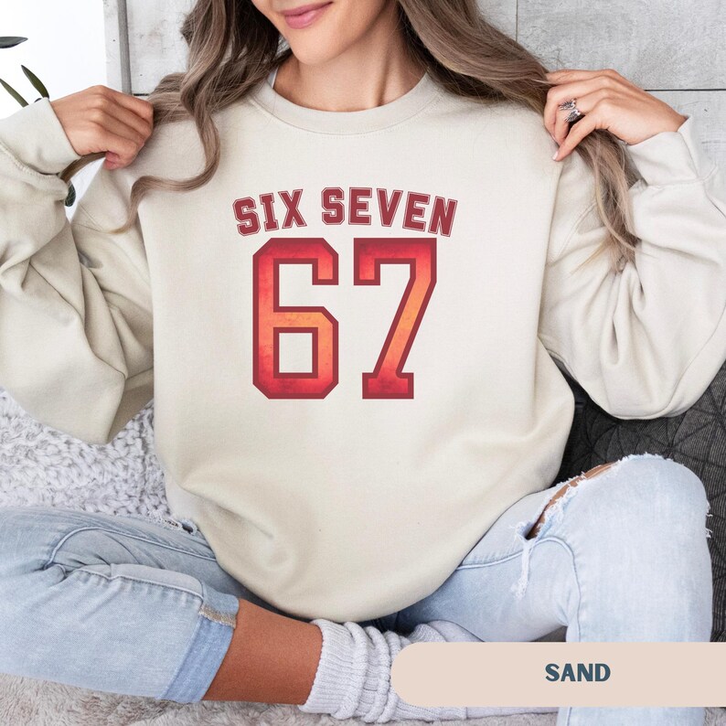 May include: A sand-colored sweatshirt with the text "SIX SEVEN" above the number "67" in a red and orange gradient. The word "SAND" is printed at the bottom of the image. The sweatshirt is a casual style.