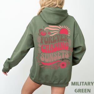 Pode incluir: Moletom verde militar com um gr&aacute;fico nas costas que diz "Forever Chasing Sunsets" em letras vermelhas e marrons. O design inclui um sol vermelho, elementos florais rosa e marrons e o texto "MILITARY GREEN" na parte inferior.