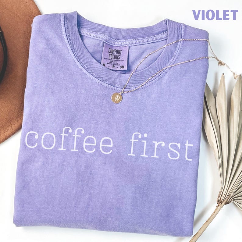 K&ouml;nnte beinhalten: Ein violettes Comfort Colors T-Shirt mit dem wei&szlig;en Schriftzug "coffee first". Eine goldene Halskette mit Anh&auml;nger liegt auf dem gefalteten Shirt. Ein brauner Hut ist teilweise sichtbar.