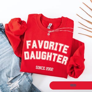 Pode incluir: Uma camisola vermelha com as palavras "FAVORITE DAUGHTER" em letras brancas, com "SINCE 2002" abaixo. Um colar de prata est&aacute; sobre a camisola. A camisola est&aacute; ao lado de um par de jeans azuis.