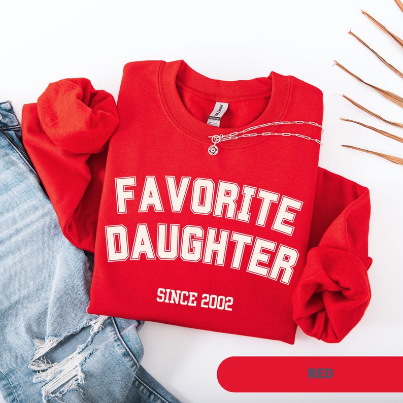 Pode incluir: Uma camisola vermelha com as palavras "FAVORITE DAUGHTER" em letras brancas, com "SINCE 2002" abaixo. Um colar de prata est&aacute; sobre a camisola. A camisola est&aacute; ao lado de um par de jeans azuis.