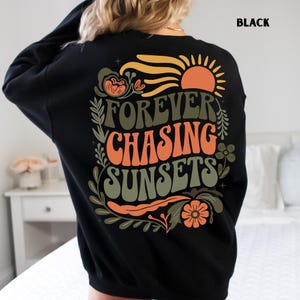 Pode incluir: Camisola preta com a frase "FOREVER CHASING SUNSETS" em laranja e verde, com um desenho de sol e flores. A palavra "BLACK" est&aacute; impressa no canto superior direito. Camisola de estilo casual.