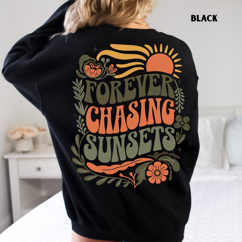 Pode incluir: Camisola preta com a frase "FOREVER CHASING SUNSETS" em laranja e verde, com um desenho de sol e flores. A palavra "BLACK" est&aacute; impressa no canto superior direito. Camisola de estilo casual.