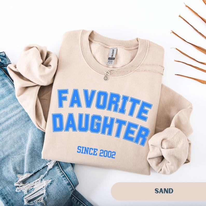 Pode incluir: Um moletom cor de areia com as palavras "FAVORITE DAUGHTER" em azul, com "SINCE 2002" abaixo. Um colar de prata com um pequeno pingente est&aacute; sobre a camisa. A palavra "SAND" est&aacute; na parte inferior.