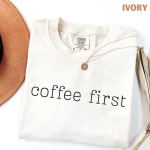 K&ouml;nnte beinhalten: Elfenbeinfarbenes T-Shirt mit dem schwarzen Schriftzug "coffee first" und einer goldenen Halskette mit Anh&auml;nger. Ein brauner Hut ist links teilweise sichtbar. Das Shirt hat ein Comfort Colors-Etikett.