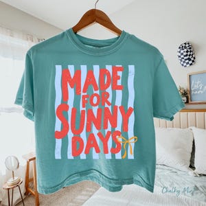Peut inclure: T-shirt de couleur sarcelle avec l'inscription "MADE FOR SUNNY DAYS" en rouge sur fond de rayures verticales bleu clair. Un n&oelig;ud jaune est en bas. Le t-shirt est suspendu &agrave; un cintre en bois.
