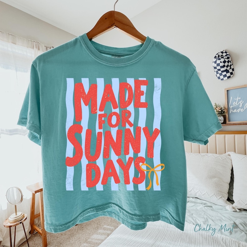 Peut inclure: T-shirt de couleur sarcelle avec l'inscription "MADE FOR SUNNY DAYS" en rouge sur fond de rayures verticales bleu clair. Un n&oelig;ud jaune est en bas. Le t-shirt est suspendu &agrave; un cintre en bois.