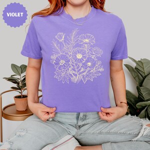K&ouml;nnte beinhalten: Ein violettes T-Shirt mit einem hellbeigen Blumendesign. Das Design umfasst verschiedene Blumen und Bl&auml;tter. Das Wort "VIOLET" befindet sich in einem violetten Kreis oben links.