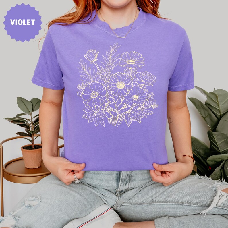 K&ouml;nnte beinhalten: Ein violettes T-Shirt mit einem hellbeigen Blumendesign. Das Design umfasst verschiedene Blumen und Bl&auml;tter. Das Wort "VIOLET" befindet sich in einem violetten Kreis oben links.