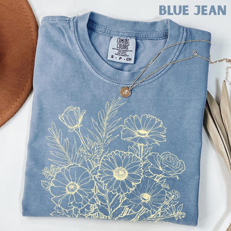 K&ouml;nnte beinhalten: Ein blaues Jeans-T-Shirt mit einem hellgelben Blumendesign. Das Shirt hat einen Rundhalsausschnitt und kurze &Auml;rmel. Eine goldene Halskette mit einem kleinen Anh&auml;nger ist &uuml;ber dem Shirt drapiert. Der Text "BLUE JEAN" steht oben.