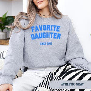 Pode incluir: Camisola cinza atl&eacute;tica com a frase "FAVORITE DAUGHTER" em letras azuis, com "SINCE 2002" por baixo. A camisola &eacute; de gola redonda e manga comprida. O canto inferior direito da camisola tem o texto "ATHLETIC GRAY".