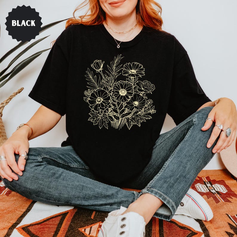 K&ouml;nnte beinhalten: Schwarzes T-Shirt mit einem goldenen Blumenstrau&szlig;-Design. Das Shirt hat kurze &Auml;rmel und einen Rundhalsausschnitt. Das Wort "BLACK" ist auf einem schwarzen Kreis oben links aufgedruckt. Das Model tr&auml;gt blaue Jeans und wei&szlig;e Turnschuhe.
