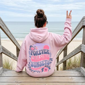 Pode incluir: Um moletom rosa claro com as palavras "FOREVER CHASING SUNSETS" em um design floral. O moletom &eacute; usado em uma escada de madeira com vista para uma praia e o oceano.