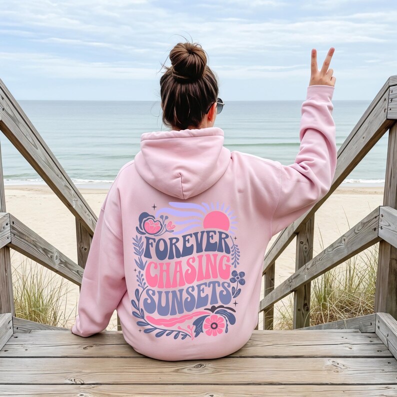 Pode incluir: Um moletom rosa claro com as palavras "FOREVER CHASING SUNSETS" em um design floral. O moletom &eacute; usado em uma escada de madeira com vista para uma praia e o oceano.