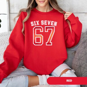 May include: A red sweatshirt with the text "SIX SEVEN" above the number "67". The numbers are white with an orange outline. The sweatshirt is a solid red color.