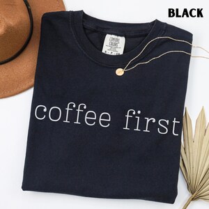 K&ouml;nnte beinhalten: Schwarzes T-Shirt mit Rundhalsausschnitt und dem wei&szlig;en Schriftzug "coffee first". Eine goldene Halskette und ein brauner Hut sind ebenfalls zu sehen. Das Shirt hat ein Etikett mit der Aufschrift "Comfort Colors".