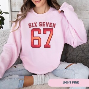 May include: Light pink sweatshirt with the text "SIX SEVEN" above the number "67" in a red and orange gradient. The sweatshirt is a solid light pink color. The text "LIGHT PINK" is at the bottom of the image.