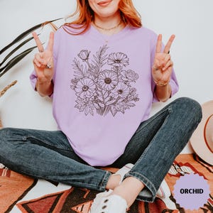 K&ouml;nnte beinhalten: Ein helllila T-Shirt mit einem schwarzen Blumenstrau&szlig;-Design. Die Grafik enth&auml;lt G&auml;nsebl&uuml;mchen, Rosen und andere botanische Elemente. Das Wort "ORCHID" ist auf einem lila Aufkleber aufgedruckt. Die Person tr&auml;gt eine blaue Jeans.