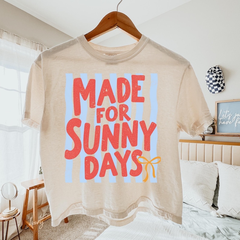 Peut inclure: T-shirt beige avec l'inscription "MADE FOR SUNNY DAYS" en lettres rouges, avec des rayures verticales bleues et un n&oelig;ud jaune. Le t-shirt est suspendu &agrave; un cintre en bois.
