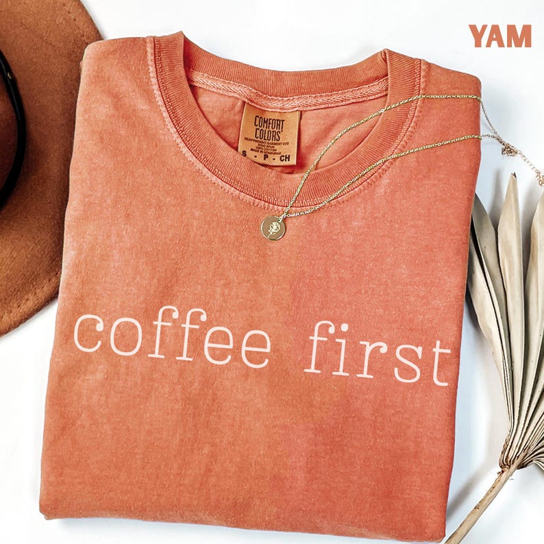 K&ouml;nnte beinhalten: Ein korallenfarbenes Comfort Colors T-Shirt mit dem wei&szlig;en Aufdruck "coffee first". Eine goldene Halskette mit Anh&auml;nger liegt auf dem Shirt. Ein brauner Hut und eine getrocknete Pflanze befinden sich im Hintergrund.