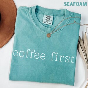K&ouml;nnte beinhalten: Meergr&uuml;nes T-Shirt mit den wei&szlig;en Worten &bdquo;coffee first&ldquo;. Eine goldene Halskette mit einem kleinen Anh&auml;nger liegt auf dem Shirt. Das Shirt ist von der Marke Comfort Colors, Gr&ouml;&szlig;e S-P-CH.