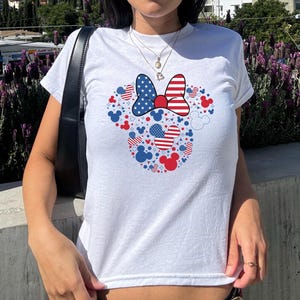May include: White t-shirt with a patriotic Minnie Mouse design. The design features a bow with a stars and stripes pattern, and red, white, and blue Mickey Mouse silhouettes and hearts. A casual, everyday wear item.