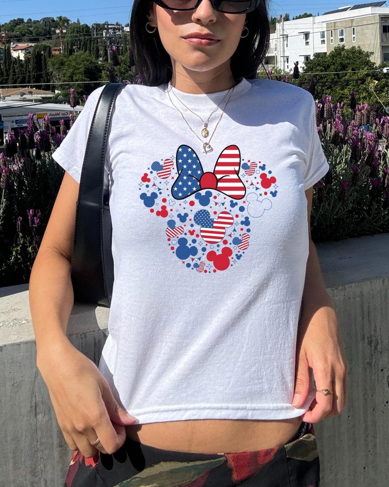 May include: White t-shirt with a patriotic Minnie Mouse design. The design features a bow with a stars and stripes pattern, and red, white, and blue Mickey Mouse silhouettes and hearts. A casual, everyday wear item.