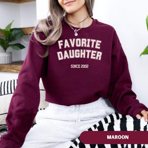 Pode incluir: Camisola bord&ocirc; com o texto "FAVORITE DAUGHTER" em letras brancas com contorno creme, e "SINCE 2002" abaixo. A camisola &eacute; de cor bord&ocirc; lisa. A palavra "MAROON" &eacute; exibida na parte inferior da imagem.
