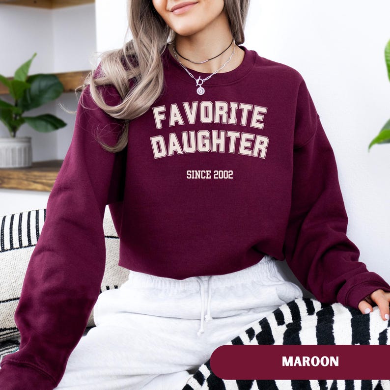 Pode incluir: Camisola bord&ocirc; com o texto "FAVORITE DAUGHTER" em letras brancas com contorno creme, e "SINCE 2002" abaixo. A camisola &eacute; de cor bord&ocirc; lisa. A palavra "MAROON" &eacute; exibida na parte inferior da imagem.