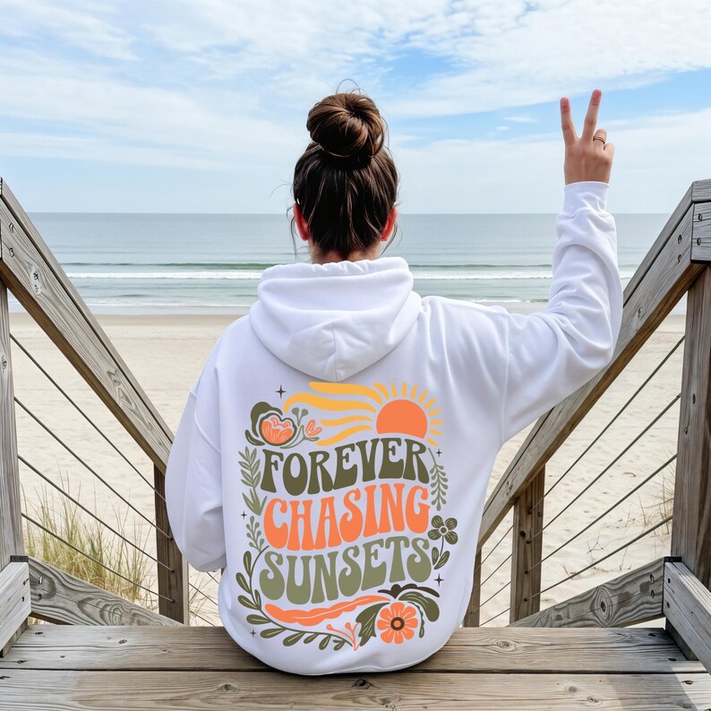 Pode incluir: Moletom branco com a frase "FOREVER CHASING SUNSETS" em laranja, verde e amarelo. O design inclui um sol, flores e folhas. A pessoa est&aacute; sentada em uma escada de madeira, com o mar ao fundo.