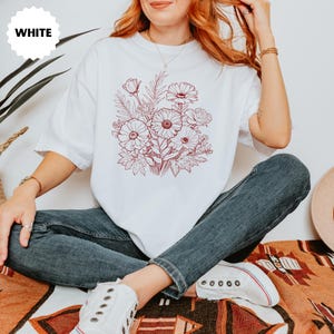 K&ouml;nnte beinhalten: Wei&szlig;es T-Shirt mit einem rotbraunen Blumendesign. Die Grafik zeigt einen Blumenstrau&szlig; mit G&auml;nsebl&uuml;mchen und Rosen. Das Wort "WHITE" befindet sich in einem wei&szlig;en Kreis oben links.