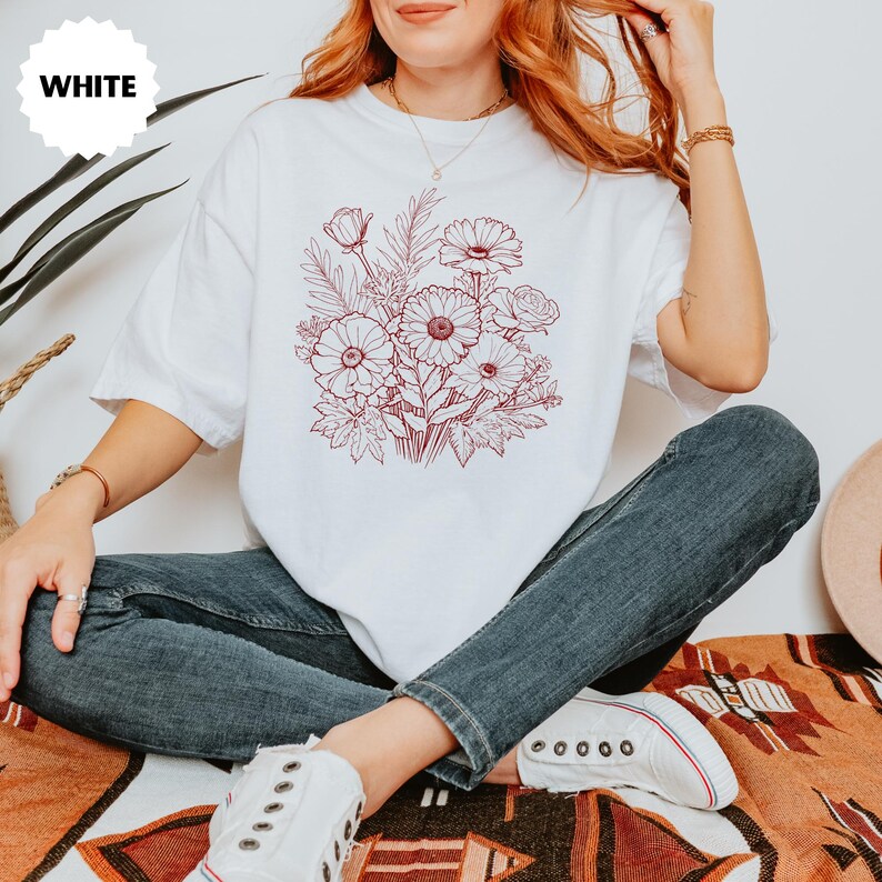K&ouml;nnte beinhalten: Wei&szlig;es T-Shirt mit einem rotbraunen Blumendesign. Die Grafik zeigt einen Blumenstrau&szlig; mit G&auml;nsebl&uuml;mchen und Rosen. Das Wort "WHITE" befindet sich in einem wei&szlig;en Kreis oben links.