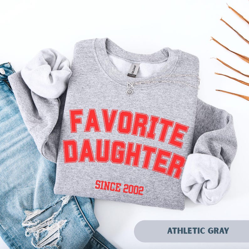 Pode incluir: Camisola cinza desportiva com as palavras "FAVORITE DAUGHTER" em letras vermelhas, com "SINCE 2002" por baixo. A camisola est&aacute; dobrada com as mangas mostrando o forro branco. Um colar de prata est&aacute; sobre a camisola.
