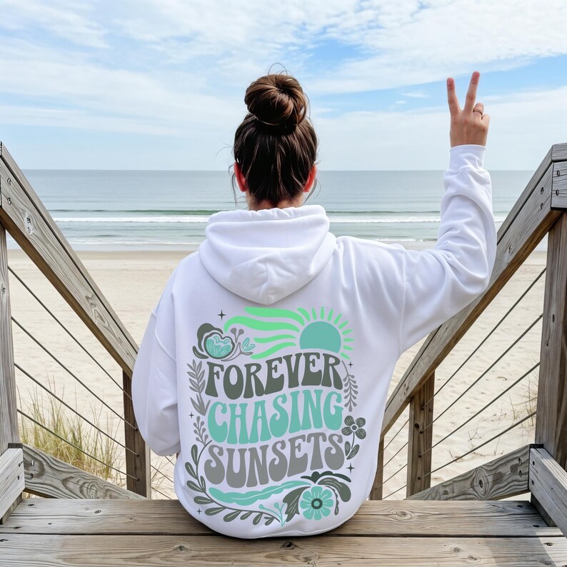 Pode incluir: Moletom branco com a frase "FOREVER CHASING SUNSETS" em verde-azulado, cinza e verde. O design inclui um sol, ondas e elementos florais. A pessoa est&aacute; sentada em degraus de madeira, com o mar ao fundo.