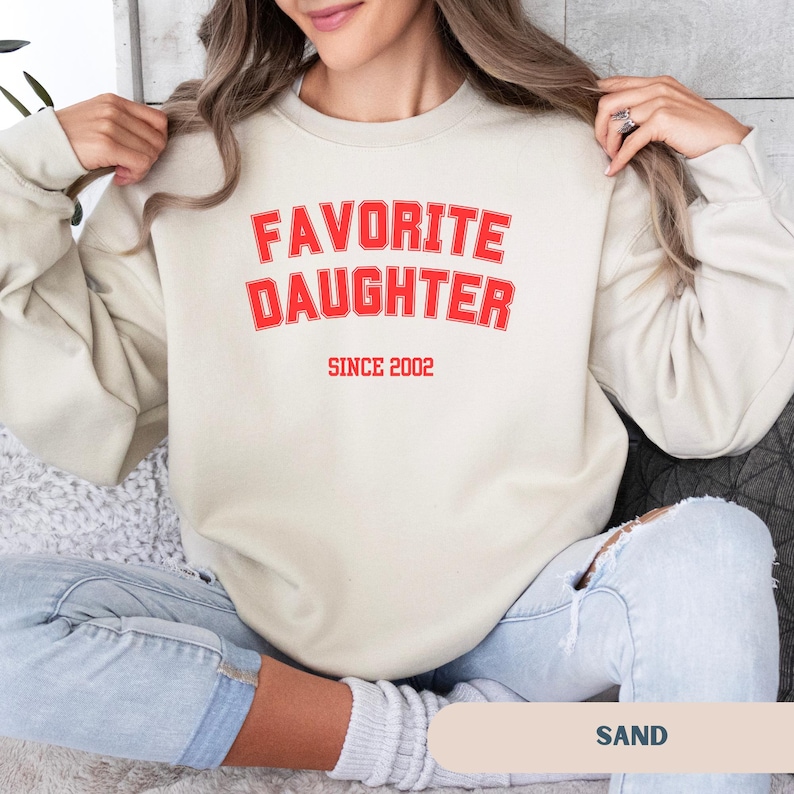 Pode incluir: Um moletom cor de areia com o texto vermelho "FAVORITE DAUGHTER" acima de "SINCE 2002". O moletom casual &eacute; um estilo confort&aacute;vel, adequado para o uso di&aacute;rio.