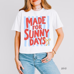 Peut inclure: T-shirt blanc avec l'inscription "MADE FOR SUNNY DAYS" en rouge, avec des rayures verticales bleues et un n&oelig;ud jaune. Le t-shirt est de style d&eacute;contract&eacute; &agrave; manches courtes.