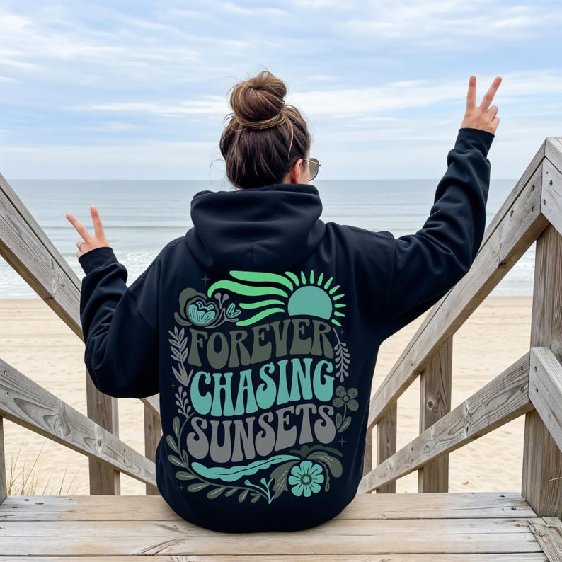 Pode incluir: Moletom preto com a frase "FOREVER CHASING SUNSETS" em uma fonte retr&ocirc;, com um design de sol e floral. O moletom &eacute; usado em uma escada de madeira que leva a uma praia com o oceano ao fundo.