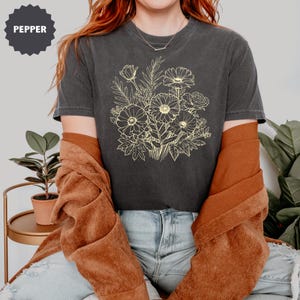 K&ouml;nnte beinhalten: Dunkelgraues T-Shirt mit einem hellgelben Blumenstrau&szlig;-Design. Der Strau&szlig; enth&auml;lt G&auml;nsebl&uuml;mchen und Rosen. Das Wort "PEPPER" befindet sich in einem dunkelgrauen Kreis oben links. Das Shirt wird mit einer rostfarbenen Jacke getragen.
