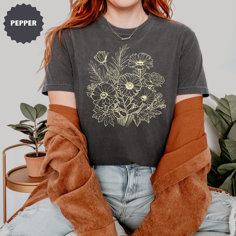 K&ouml;nnte beinhalten: Dunkelgraues T-Shirt mit einem hellgelben Blumenstrau&szlig;-Design. Der Strau&szlig; enth&auml;lt G&auml;nsebl&uuml;mchen und Rosen. Das Wort "PEPPER" befindet sich in einem dunkelgrauen Kreis oben links. Das Shirt wird mit einer rostfarbenen Jacke getragen.