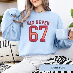 May include: Light blue sweatshirt with the text "SIX SEVEN" above the number "67" in a red and orange collegiate style font. The sweatshirt is a solid light blue color. A light blue mug is held in the right hand.