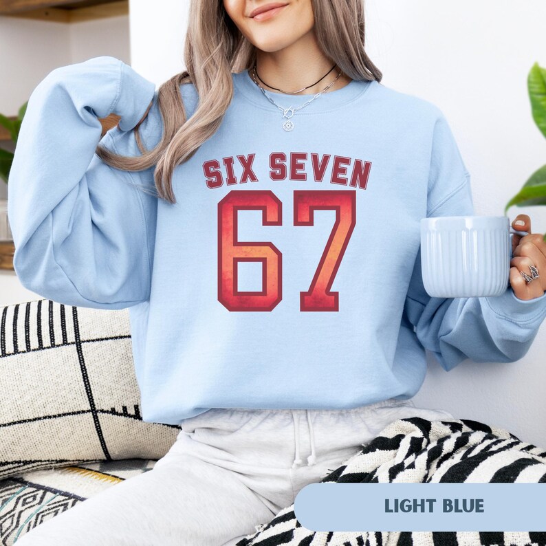 May include: Light blue sweatshirt with the text "SIX SEVEN" above the number "67" in a red and orange collegiate style font. The sweatshirt is a solid light blue color. A light blue mug is held in the right hand.