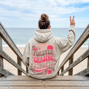 Pode incluir: Um moletom bege com o texto "FOREVER CHASING SUNSETS" em rosa e marrom, com um gr&aacute;fico de p&ocirc;r do sol. O moletom &eacute; usado por uma pessoa sentada em degraus de madeira, com o oceano ao fundo.