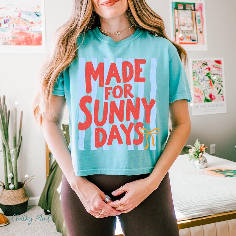 Peut inclure: Un t-shirt turquoise avec le texte "MADE FOR SUNNY DAYS" en rouge, avec des rayures verticales bleu clair. Un motif de n&oelig;ud jaune est en bas &agrave; droite. Le t-shirt est de style court.