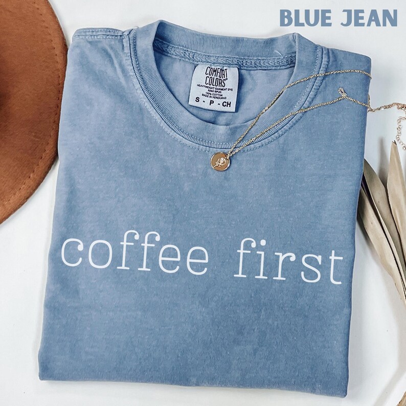 K&ouml;nnte beinhalten: Ein blaues Jeans-T-Shirt mit den wei&szlig;en Worten "coffee first" aufgedruckt. Das Shirt hat einen Rundhalsausschnitt und kurze &Auml;rmel. Eine goldene Halskette mit Anh&auml;nger ist &uuml;ber dem Shirt drapiert.