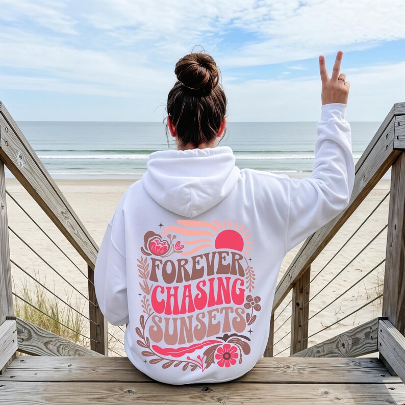 Pode incluir: Moletom branco com a frase "FOREVER CHASING SUNSETS" em um design retr&ocirc;. O design inclui um sol rosa, flores e um esquema de cores de p&ocirc;r do sol. O moletom &eacute; usado em um deck de madeira com vista para o oceano.